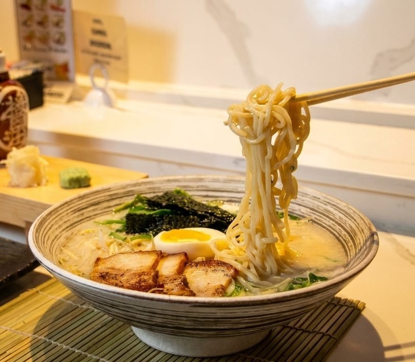 Chopsticks lifting noodles from a bowl of ramen with pork, egg, and seaweed.